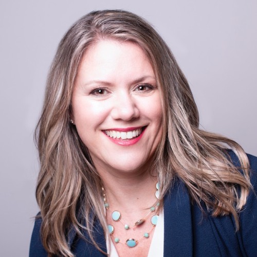 Smiling woman with long blonde hair, wearing a blue blazer and turquoise necklace, on a light background.
