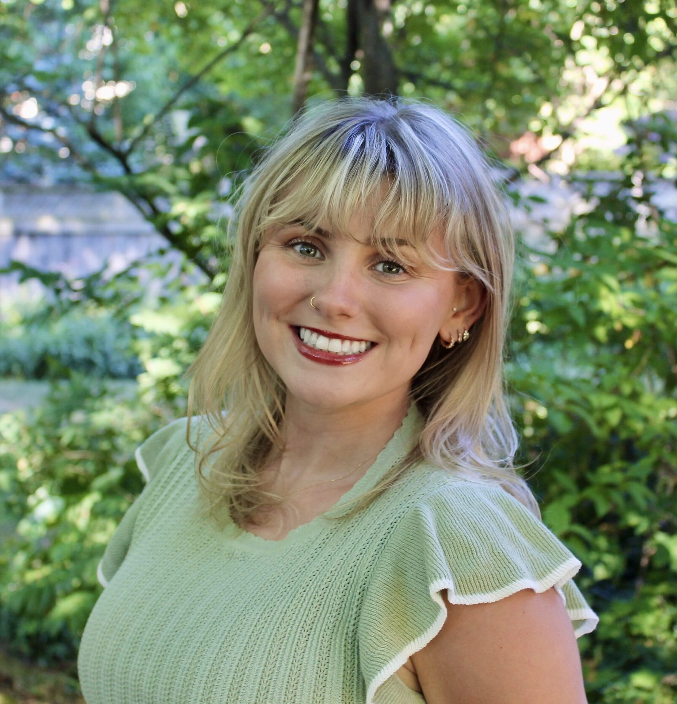 A woman with blonde hair and a green top smiles outdoors in a lush, leafy garden.