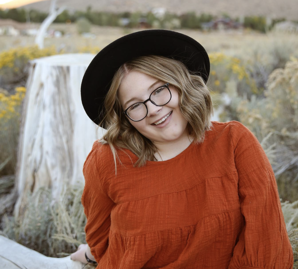 Person wearing glasses and a black hat, smiling outdoors with grass and a tree stump in the background.