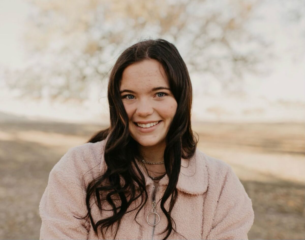Smiling person with long dark hair wearing a pink fuzzy jacket outdoors, with trees blurred in the background.