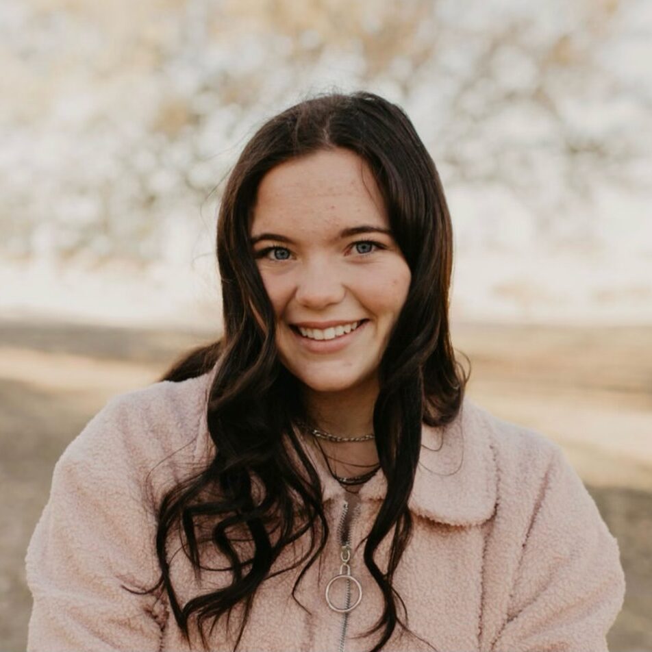 Smiling person with long dark hair wearing a pink fuzzy jacket outdoors, with trees blurred in the background.