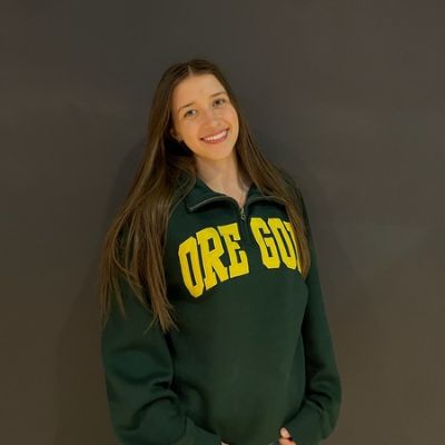 Woman smiling, wearing a dark green sweatshirt with yellow OREGON lettering, against a plain dark background.
