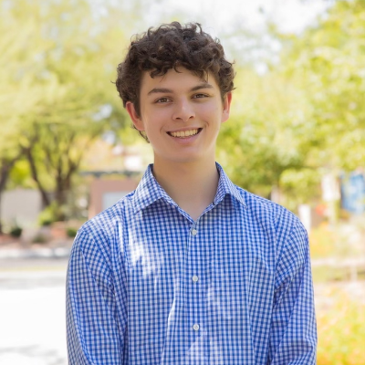 A person with curly hair in a blue checkered shirt smiles outdoors with trees in the background.