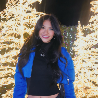 A woman in a blue jacket smiles in front of illuminated holiday trees at night.