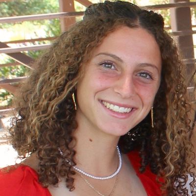 Smiling woman with curly hair in a red top, wearing pearl necklace and earrings, outdoors under a pergola.