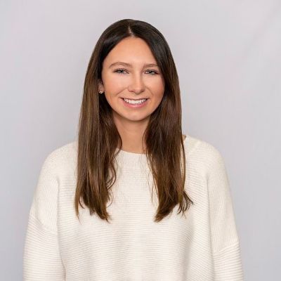 Woman with long brown hair in a white sweater smiling in front of a plain, light gray background.