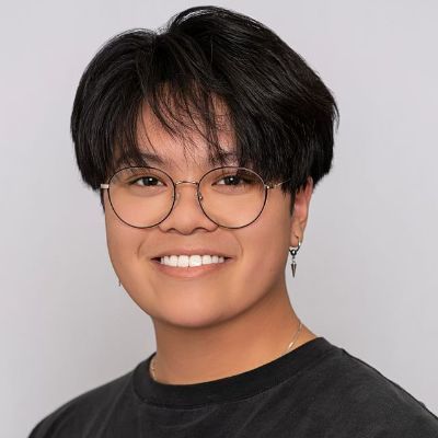 Person with short dark hair and round glasses smiling against a gray background.