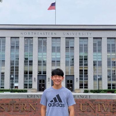 A person stands smiling in front of Northeastern University with the American flag flying above.
