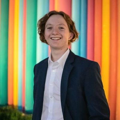 Person smiling in front of colorful, vertical lights, wearing a white shirt and dark blazer.