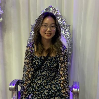 A woman in a floral dress sits on a silver ornate chair against a white curtain backdrop.