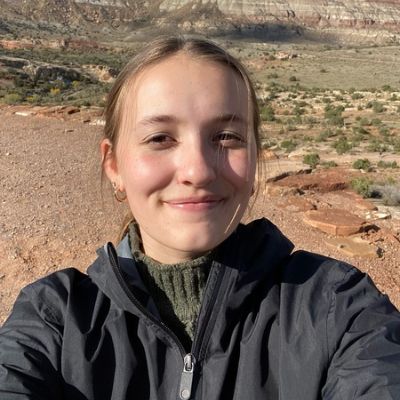 A person smiling outdoors with a rocky landscape and clear sky in the background.