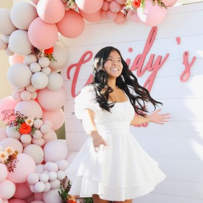 Woman in a white dress stands joyfully in front of a pink balloon arch and a sign.