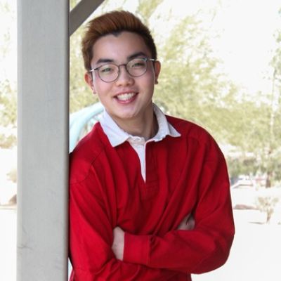 A person in a red sweater, smiling and leaning against a column with trees in the background.