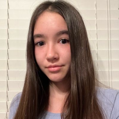 A person with long brown hair and a light blue shirt stands in front of white blinds, looking at the camera.
