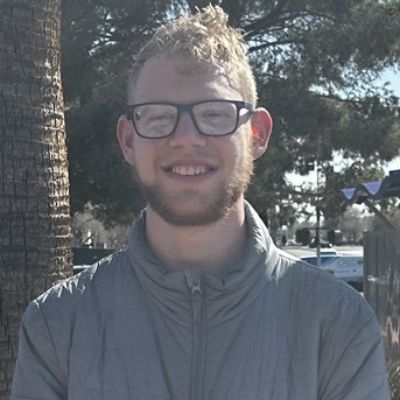 A smiling person with glasses and a gray jacket stands outdoors near a tree on a sunny day.