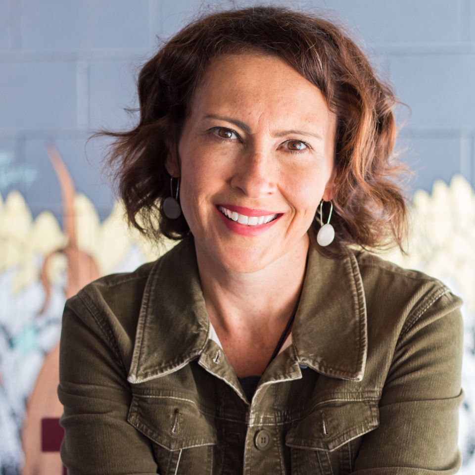 Smiling woman with curly hair, wearing a green jacket and earrings, in front of a mural background.