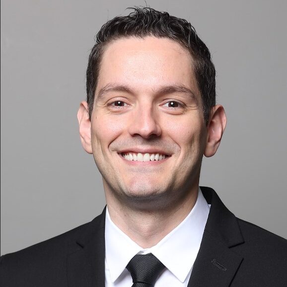 Man in a black suit and tie smiling against a gray background.
