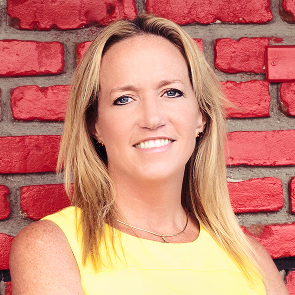 Smiling person with long hair in a yellow top in front of a red brick wall.