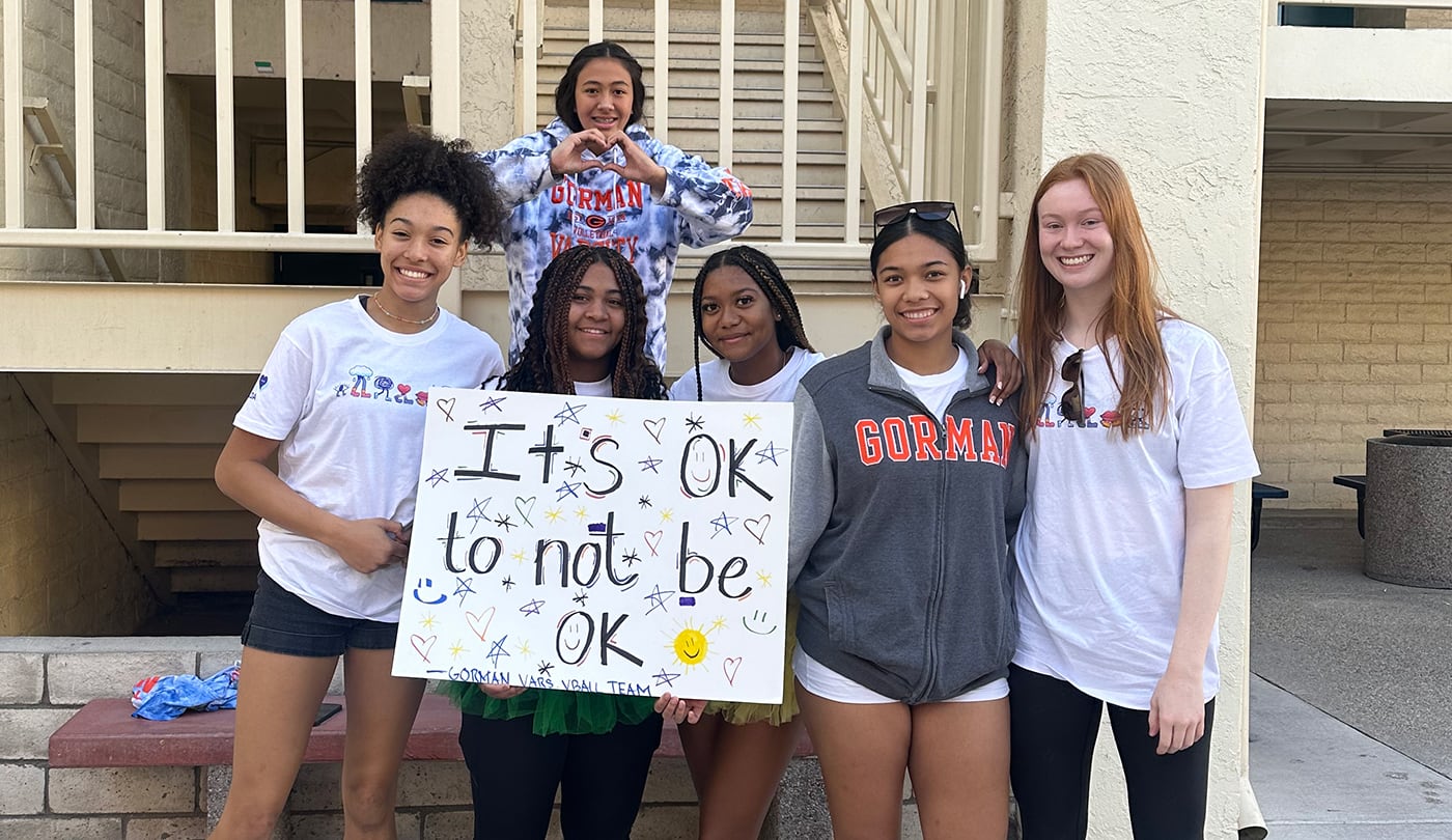 Six smiling people with a sign saying Its OK to not be OK standing outside a building.