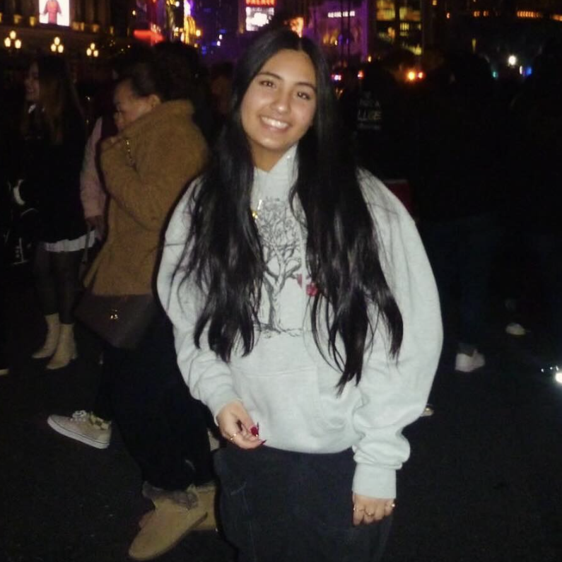 Person in a grey hoodie smiles while standing in a busy nighttime city scene with colorful lights.