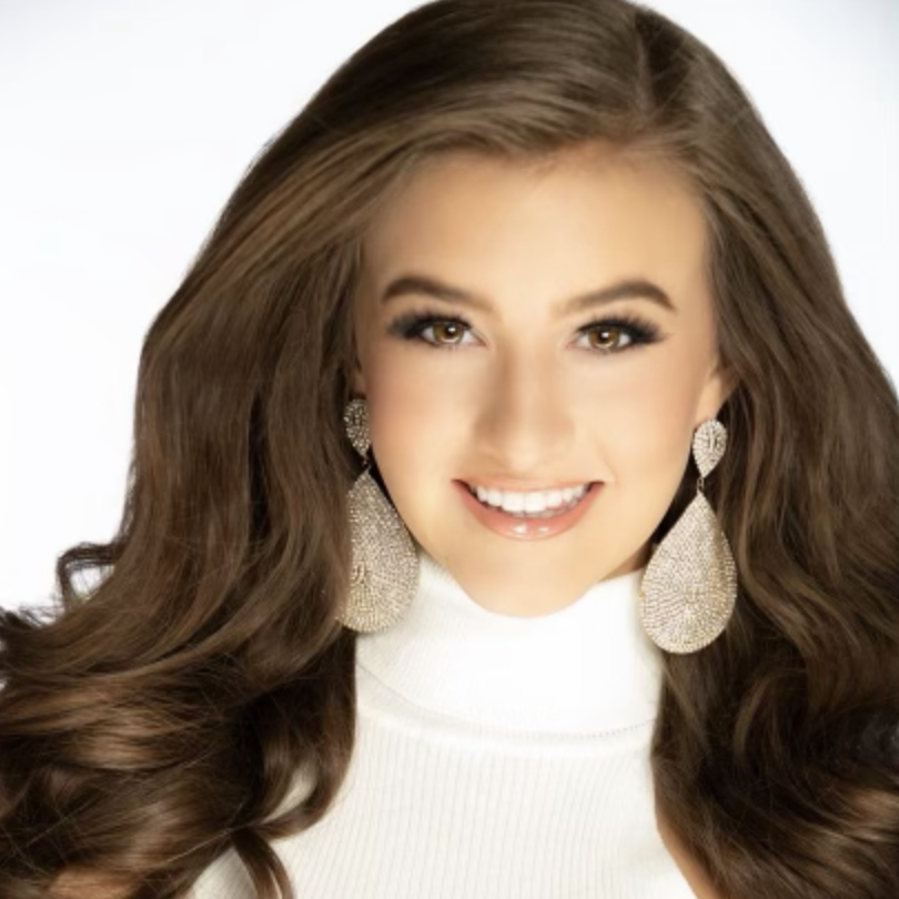 Smiling woman with long brown hair, wearing large sparkly earrings and a white turtleneck.