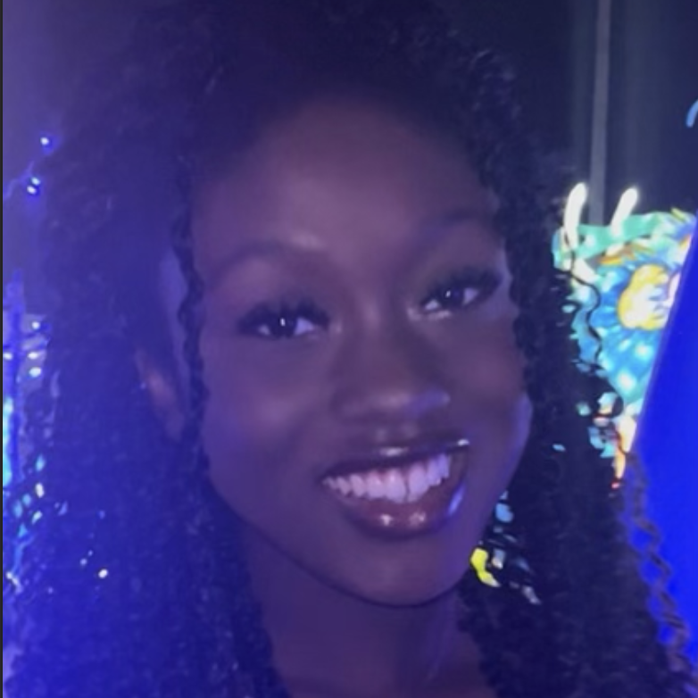 Woman smiling brightly with curly hair, standing in front of colorful, illuminated decor at night.