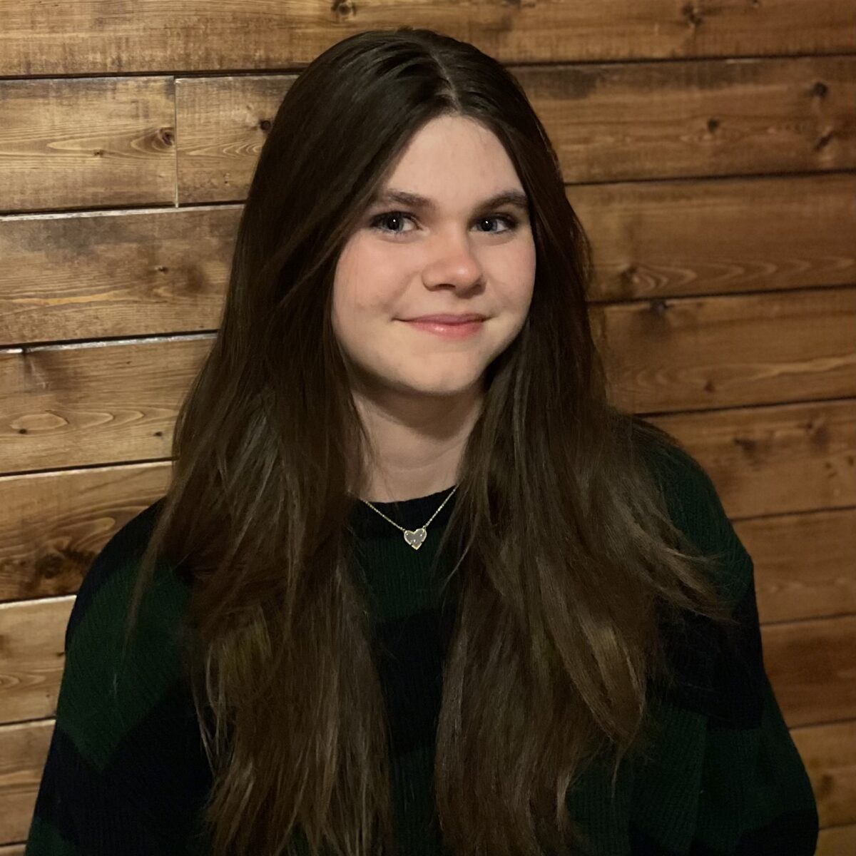 Girl with long brown hair smiling in front of a wooden wall.