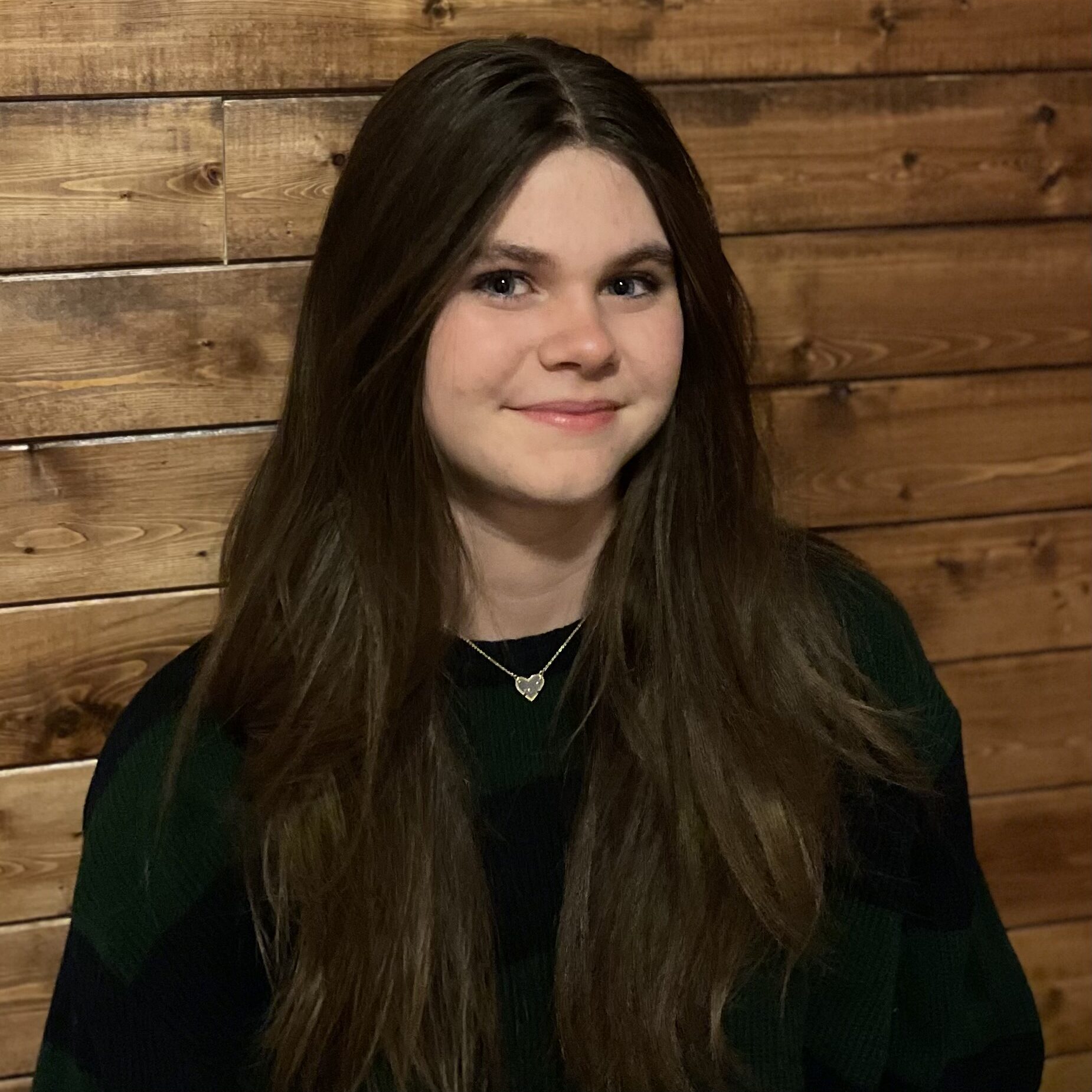 Girl with long brown hair smiling in front of a wooden wall.