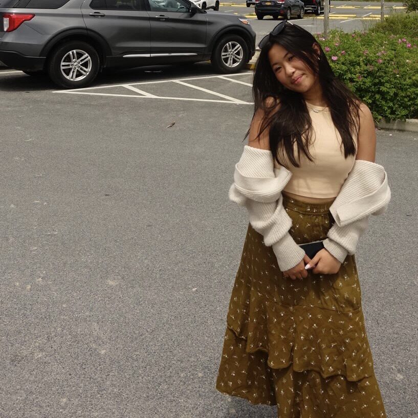 Woman in a beige top and green skirt stands smiling in a parking lot, holding a white sweater over her shoulders.