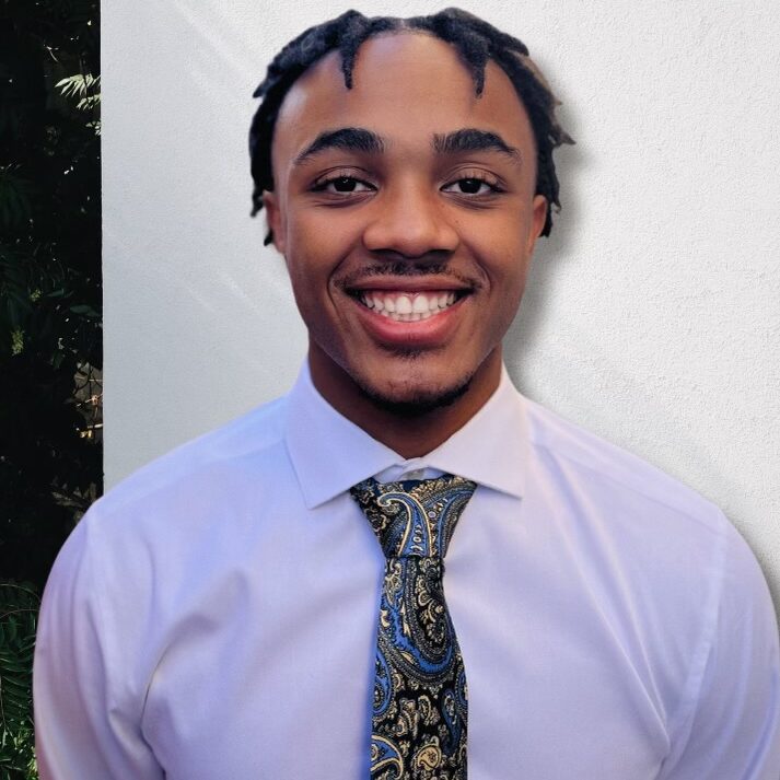 Smiling person in a white shirt and paisley tie, standing against a plain white wall.