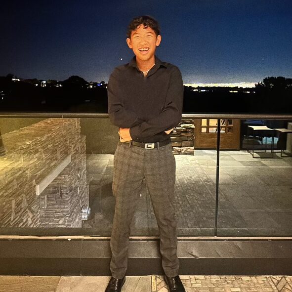 Smiling person in dark shirt and plaid pants stands on a lit patio at night with a cityscape in the background.