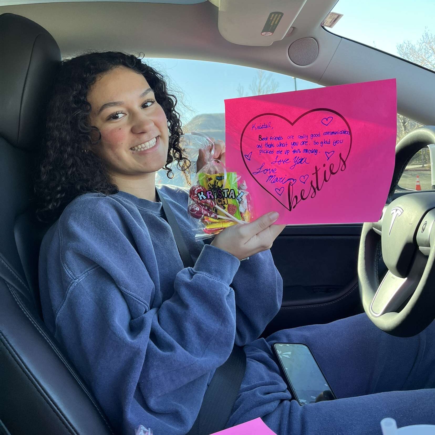 A person in a car holds a colorful card and candy bag, smiling at the camera.