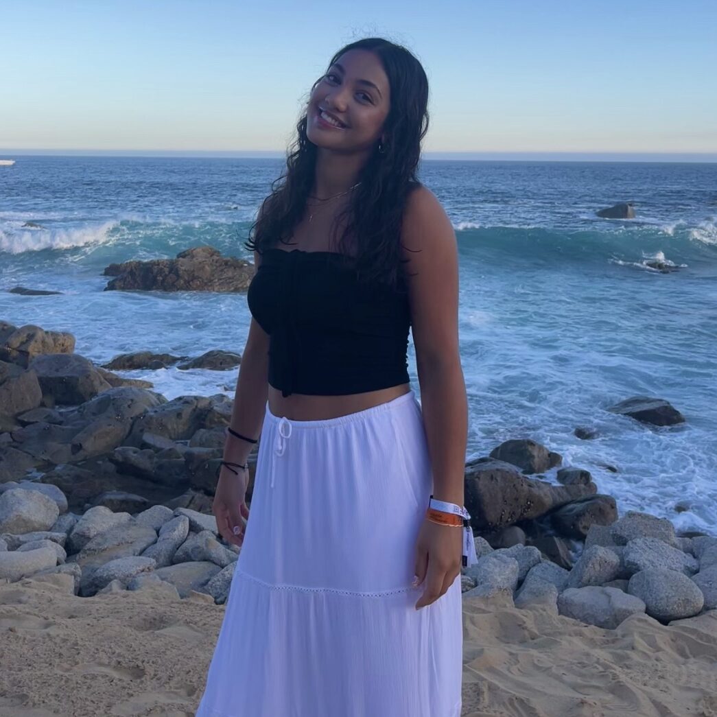 Woman smiling by the ocean, wearing a black top and white skirt, standing on a rocky beach under a clear sky.