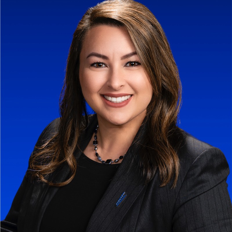 Smiling person with long hair in a black suit against a blue background.