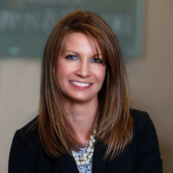 A woman with long straight hair smiling, wearing a dark blazer and a pearl necklace.