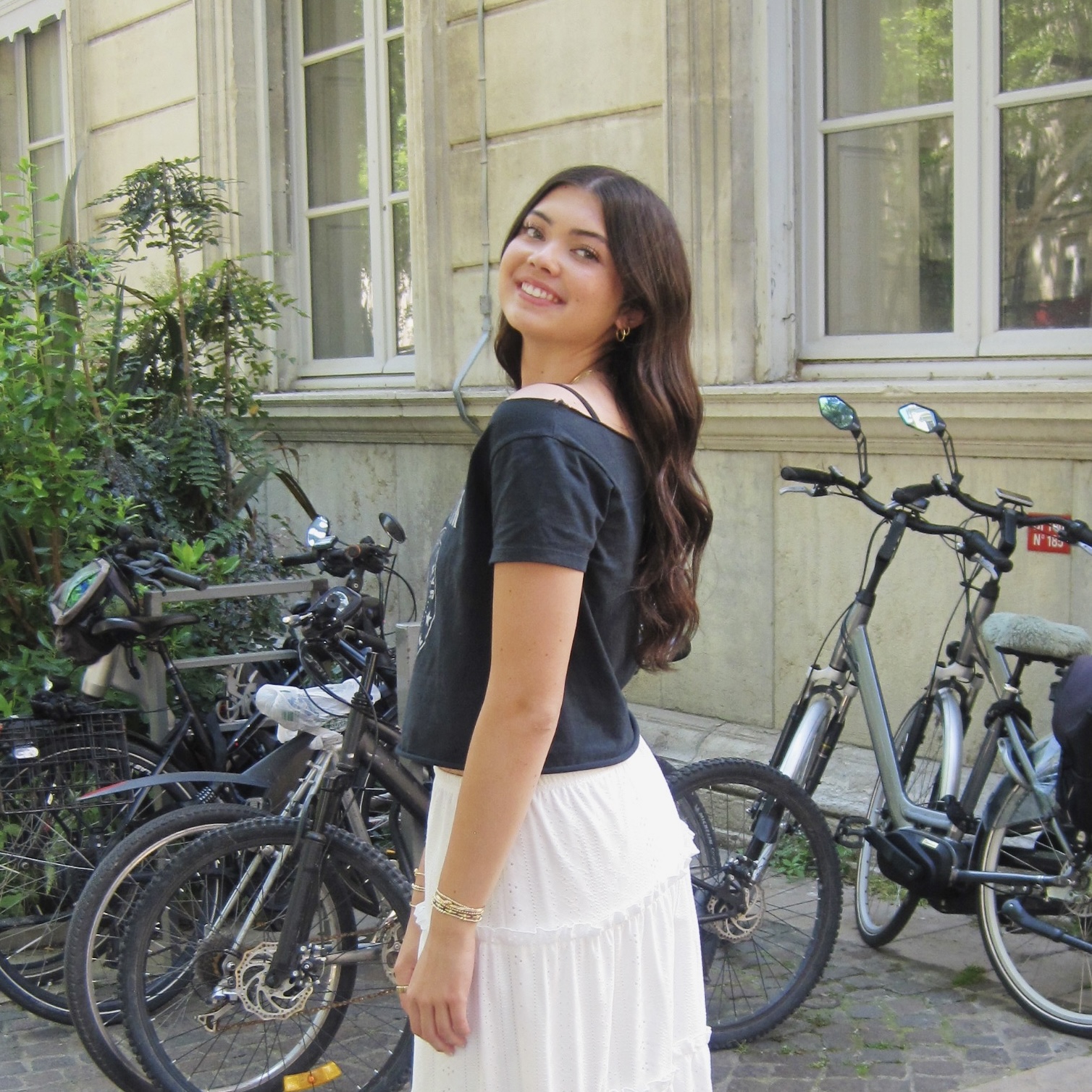 Woman with long dark hair smiles outdoors near parked bicycles and greenery, wearing a black top and white skirt.