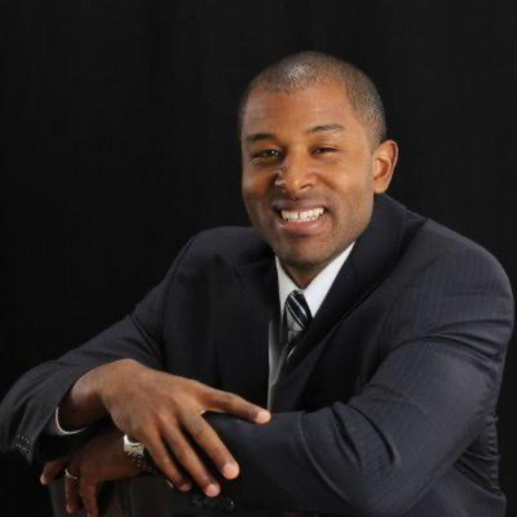 Smiling man in a dark suit sitting with arms resting on a chair, against a black background.