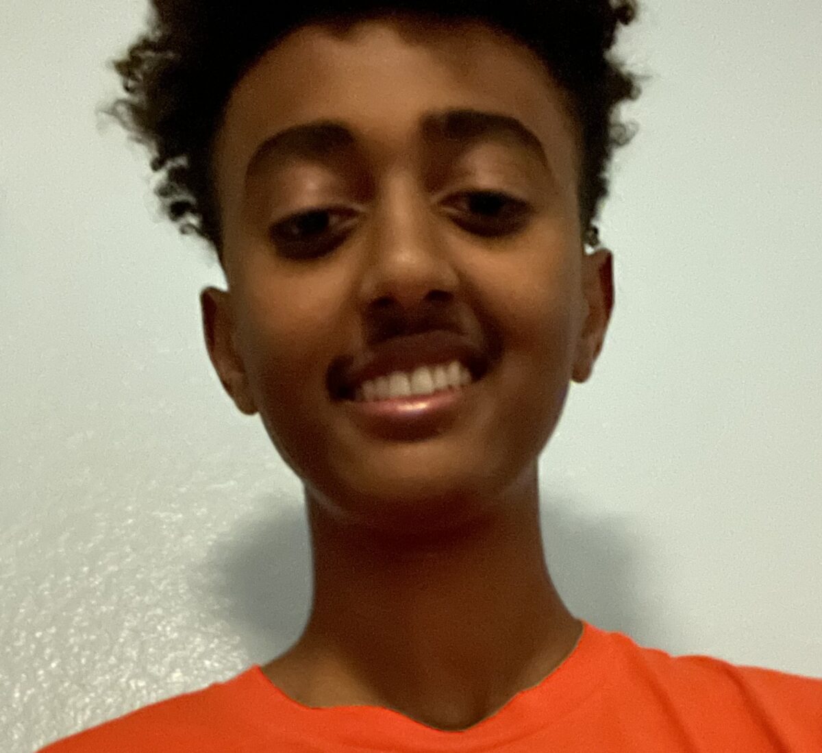 Smiling young person with short curly hair wearing an orange shirt in front of a light-colored wall.