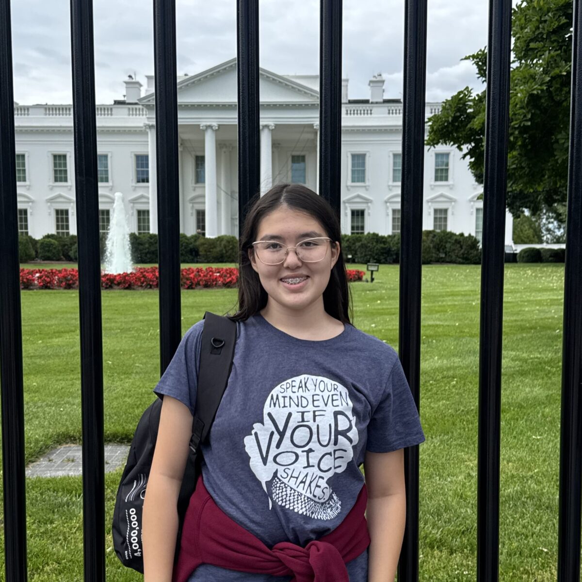 A girl stands smiling in front of the White House, behind a black iron fence on a cloudy day.
