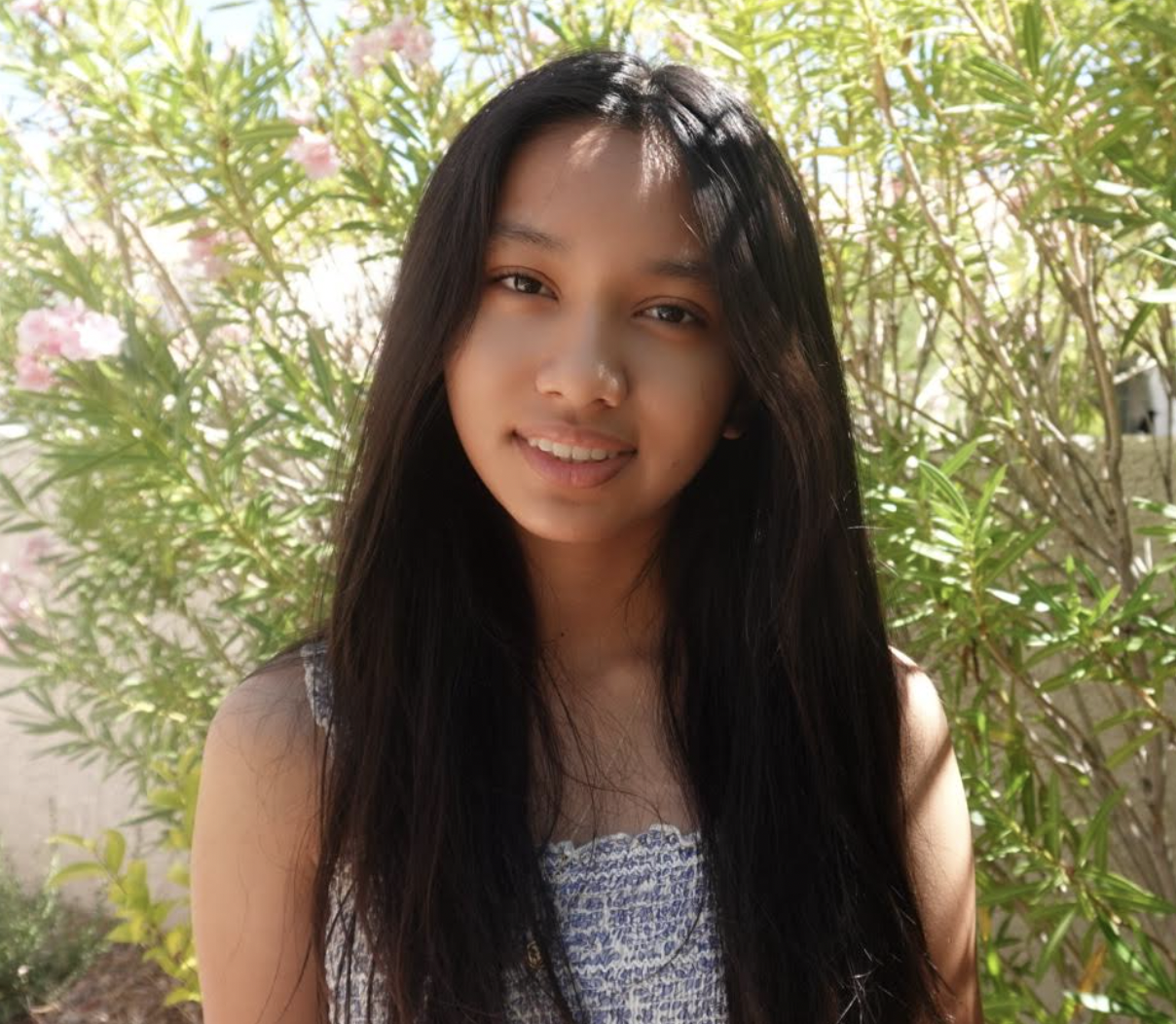 A young woman with long dark hair smiles outdoors, standing in front of green bushes and pink flowers.